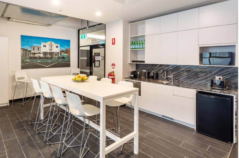 Modern office kitchen with white cupboards, long dining table and chairs, appliances, water bottles on shelves, and a wall photo of a building at spaces@115.