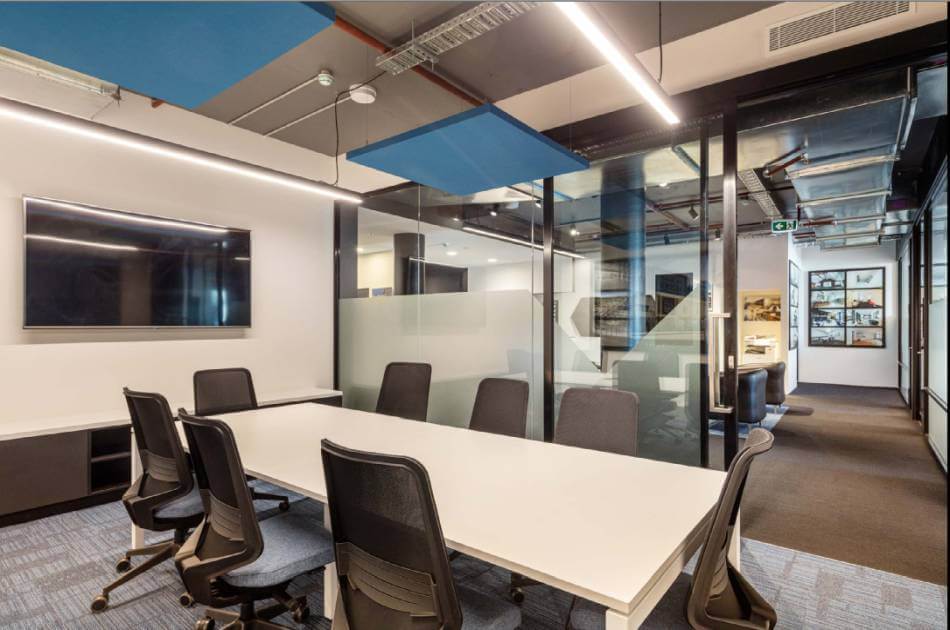 Modern conference room at spaces@115 with a long white table, black office chairs, a wall-mounted screen, glass walls, and overhead lighting. A corridor with framed pictures is visible through the glass.